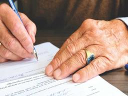 mãos de um homem mais velho assinando um documento. o homem usa um anel dourado com uma pedra azul no dedo anelar e veste uma blusa marrom.
