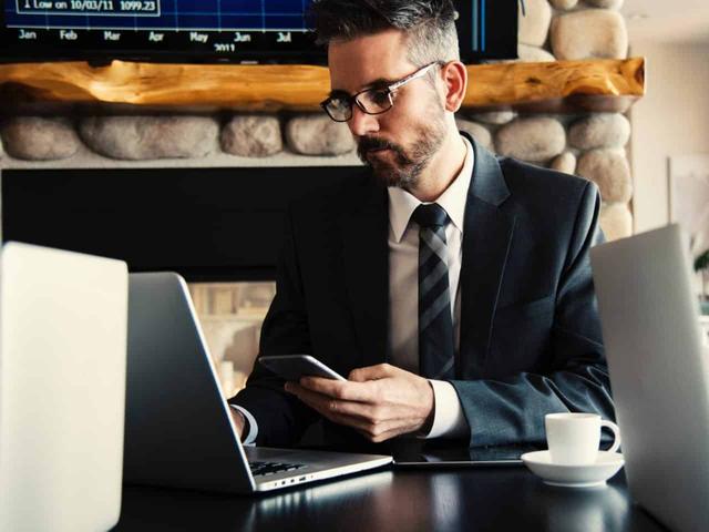 imagem de um homem com um celular na mão e olhando um computador para escolher o melhor banco para abrir conta MEI