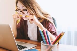 menina de óculos com um lápis na boca e fazendo expressão de dificuldade olhando para um computador. Ela está sentada em uma mesa com lápis ao seu lado