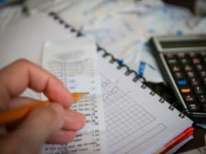 mão segurando lápis fazendo anotações em nota fiscal, com caderno e calculadora ao fundo epresentando famílias com dívidas