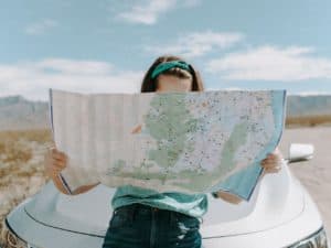 Imagem de uma mulher apoiando em um carro e observando um mapa, representando o conteúdo sobre viagens no contexto pós vacinação