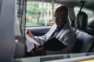 Homem negro, de terno, sentado no banco de trás de um carro, lendo alguns documentos dentro de uma pasta