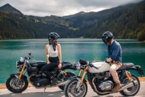 Duas motos, uma com um homem e outra com uma mulher. Ao fundo, um lago azul e montanhas. O céu está pouco nublado.