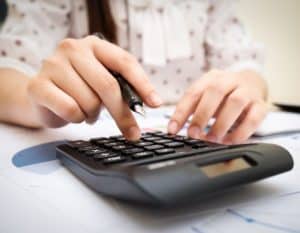 Mulher fazendo cálculos numa calculadora preta apoiada sobre uma mesa com vários papéis.
