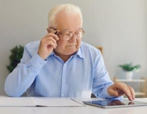 Homem idoso veste camisa social azul bebê, tem cabelos brancos, usa óculos e está olhando um tablet que está sob uma mesa branca.