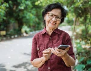 Mulher idosa veste camisa cor vinho, usa óculos e está sorrindo com um celular na mão. Ela se encontra numa rua com árvores ao fundo.
