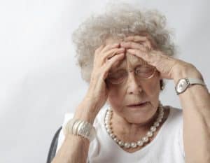 Uma mulher idosa de cabelos brancos, veste uma blusa branca, cordão de pérolas, pulseira, relógio e óculos. Está com uma expressão de preocupada, com as mãos sobre a cabeça.