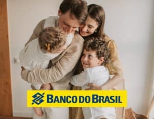 Foto de uma família se abraçando. A mãe está na direita da foto com as mãos sobre o seu filho pequeno, o pai ao lado esquerdo com uma criança menina no colo e todos sorriem. Há a logo do Banco do Brasil em amarelo e azul.