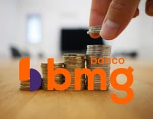Mão empilhando moedas sobre uma mesa de madeira. Há na foto a logo do banco BMG em laranja.