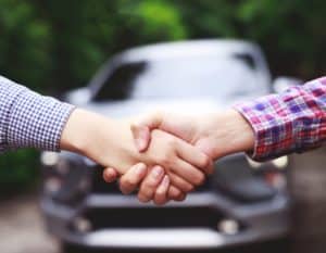 Aperto de mãos entre duas pessoas. No fundo da foto há um carro cinza.