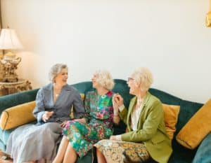 Três mulheres idosas estão sentadas num sofá rindo, olhando uma para a outra. A da esquerda veste um vestido cinza, a do meio, um vestido verde e, a da direita, um branco. Todas têm cabelos brancos.