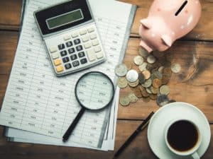 Imagem de uma folha com números, uma calculadora, uma xícara de café, uma lupa e um porquinho com algumas moedas. Foto utilizada para ilustrar uma notícia sobre a declaração retificadora IR.
