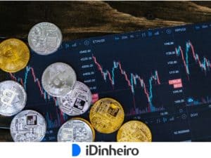 tela de computador com gráficos do mercado financeiro e, em sua frente, moedas pratas e douradas.