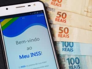 Imagem de um celular com o aplicativo Meu INSS e duas notas de 100 reais e duas de 50 reais. Imagem utilizada para ilustrar notícia sobre os pagamentos do INSS em junho.