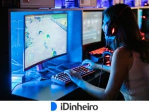 uma pessoa de camiseta e cabelo comprido com fones de ouvido, em frente a um PC gamer com fundo de led azul, segurando um teclado com luzes, com expressão de sorriso. Na tela do computador, um jogo.