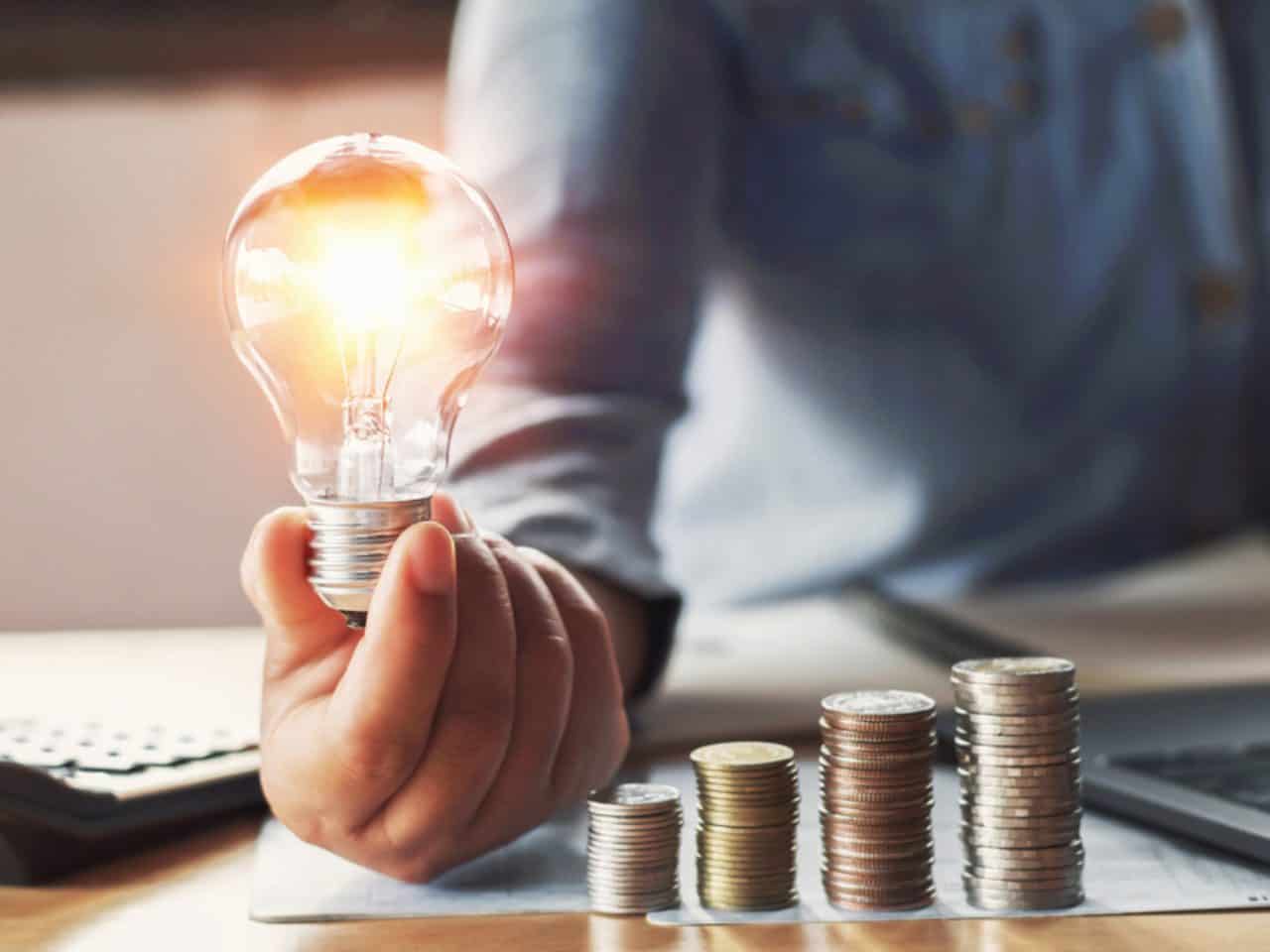 Imagem de uma pessoa segurando uma lâmpada ao lado de algumas moedas. Foto utilizada para ilustrar uma notícia sobre os impactos da redução icms energia.