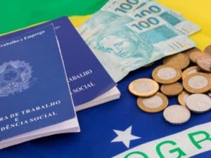 Imagem de uma carteira de trabalho, junto com uma bandeira do Brasil e algumas cédulas de 100 reais e moedas de 1 real. Foto utilizada para ilustrar uma notícia sobre o salário mínimo 2023.