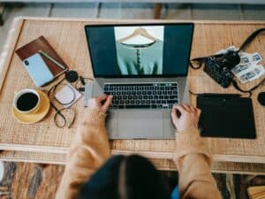 empreendedorismo digital: pessoa trabalhando em seu laptop que está sobre uma mesa de madeira juntamente de outros objetos, como uma xícara de café e seu celular.