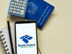Imagem de um celular com a logo da Receita Federal ao lado de uma calculadora e de uma carteira de trabalho. Foto utilizada para ilustrar uma notícia sobre a declaração pré-preenchida. Crédito: Shutterstock