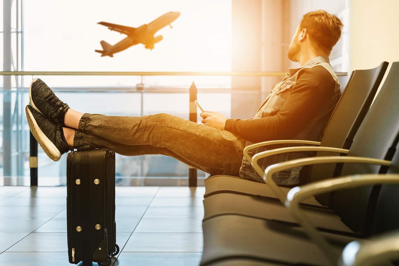 pessoa em uma poltrona de aeroporto, inclinada, relaxando com os pés sobre uma mala, observando um avião passando lá fora