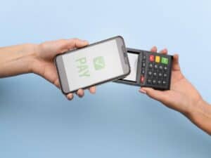 Uma mão segura um celular, enquanto outra segura uma maquininha. Ambas se encontram para finalizar um pagamento representando fluxo de dinheiro no varejo.
