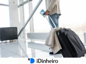 Homem em um aeroporto segurando uma passagem aérea e uma mala de rodinhas preta, com um casaco bege dobrado sobre a mala. Ele está usando calça jeans e um blazer claro. A cena sugere que ele está prestes a viajar. Ao fundo, há grandes janelas de vidro com vista para o exterior. A imagem está relacionada ao uso de benefícios de viagens, como os oferecidos pelo cartão de crédito Visa Infinite.