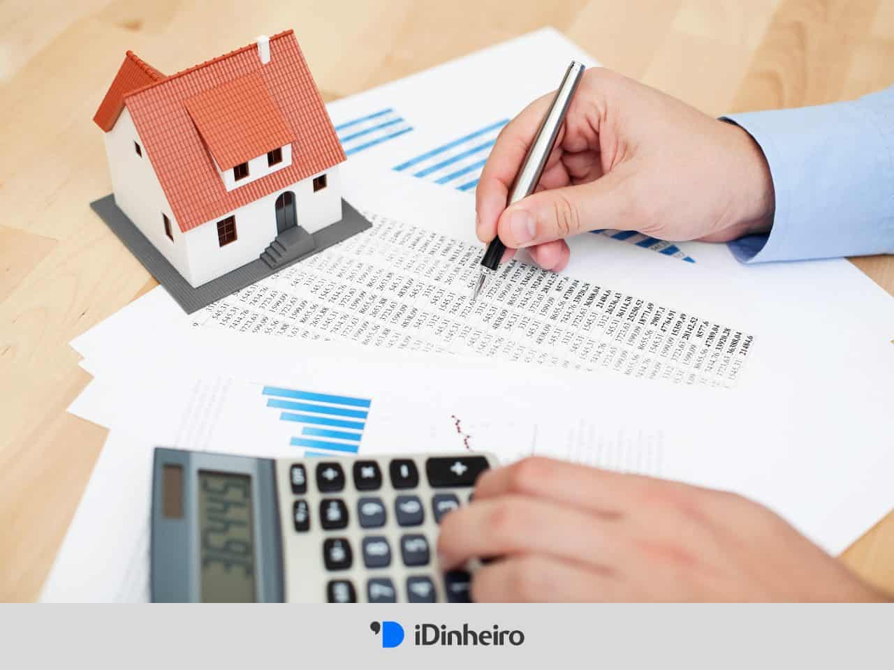 sobre uma mesa, papeis, calculadora e uma casa em miniatura. Uma mão segura uma caneta, enquanto a outra utiliza a calculadora, simbolizando o cálculo do financiamento tipo Price.